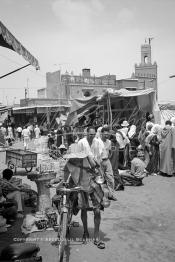 Image du Maroc Professionnelle de  Avec un chapeau de paille sur la tête et poussant son vélo, le traditionnelle vendeur et porteur d'eau dit –Guerrab- n'est pas aussi visible que celui qui est folklorique doté d'un chapeau multicolore, et d'une tenue , chargé de coupelles en cuivres ou de fer blanc qui brille sous les rayons du soleil. Cet authentique vporteur d'eau qui désaltère ceux qui ont soif, traverse l'espace de la Place Jemaa El Fana et ne fait aucunement pas partie des attractions touristiques, au fond le minaret de la Mosquée Kharbouche, Jeudi 19 Mai 1988. (Photo / Abdeljalil Bounhar)